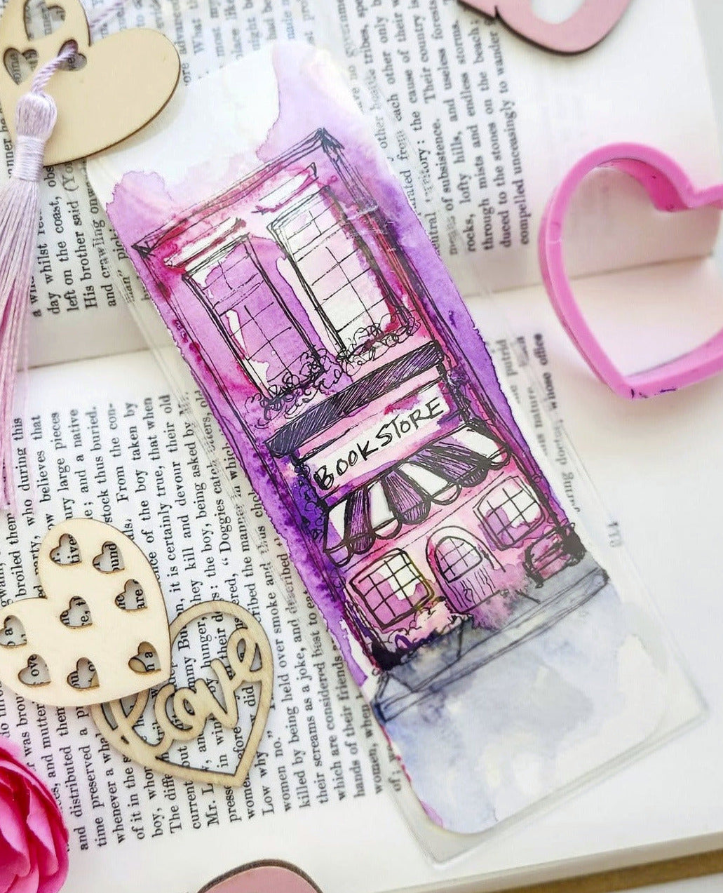 A Valentine's Day themed decorative tube featuring a pink and purple watercolor illustration of a bakery, styled on top of an open book with pink wooden XOXO letters, heart-shaped cutouts, a pink rose, and a tassel.