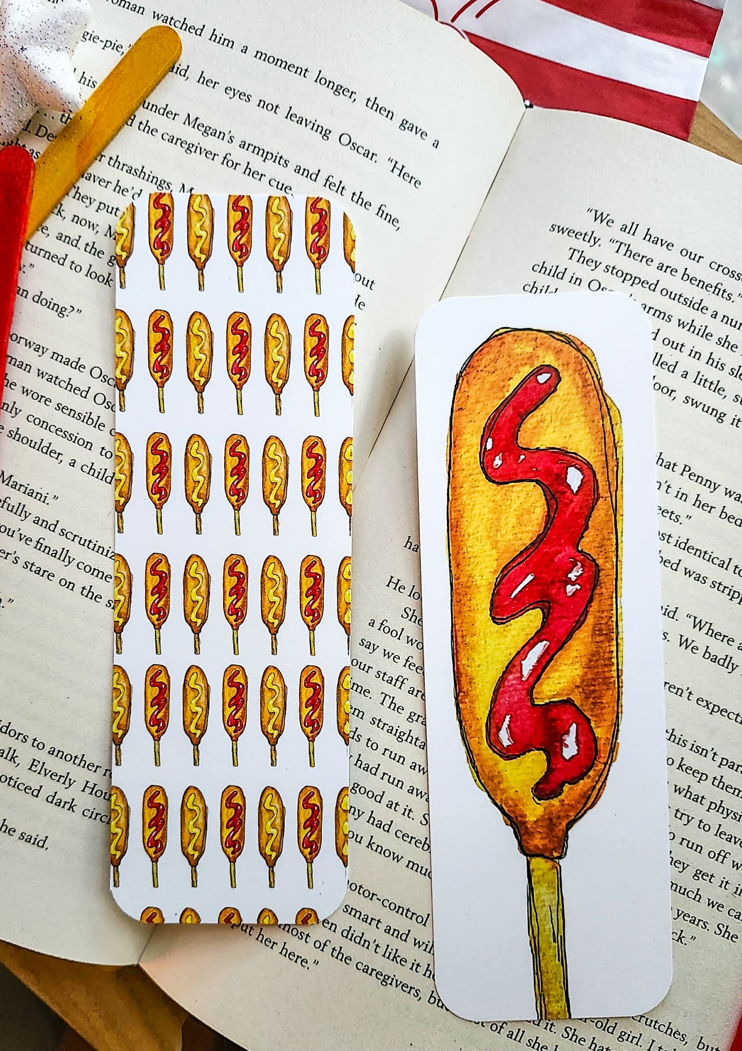 A hand-drawn illustrated bookmark shaped like a corndog with red ketchup swirl, placed on an open book next to another bookmark featuring multiple corndogs with ketchup and mustard