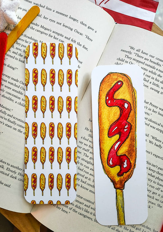 A hand-drawn illustrated bookmark shaped like a corndog with red ketchup swirl, placed on an open book next to another bookmark featuring multiple corndogs with ketchup and mustard
