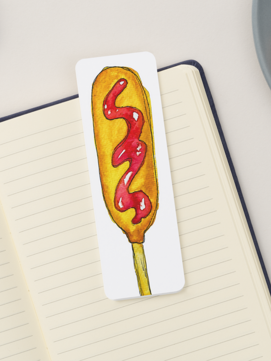 A white rectangular bookmark featuring a watercolor illustration of a hot dog with red ketchup, placed on an open lined notebook.