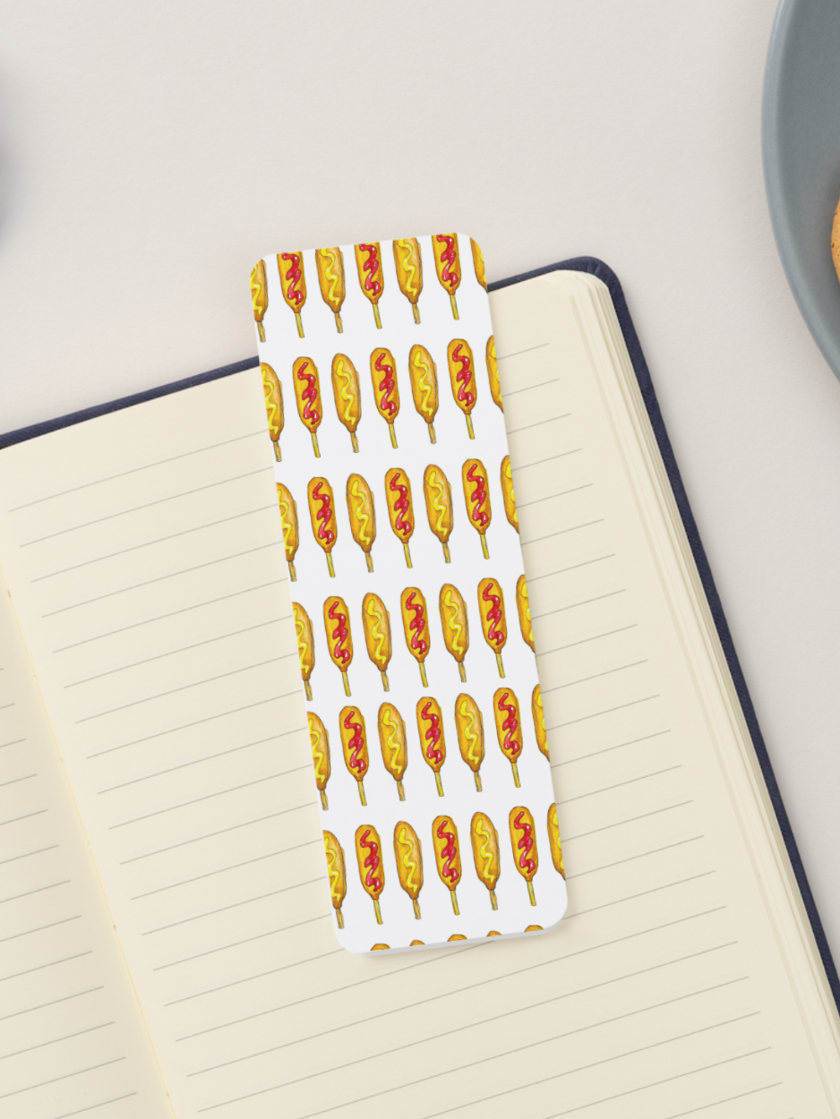 A white rectangular bookmark with a repeating pattern of illustrated corn dogs in yellow, orange, and red, placed across an open lined notebook on a light surface.