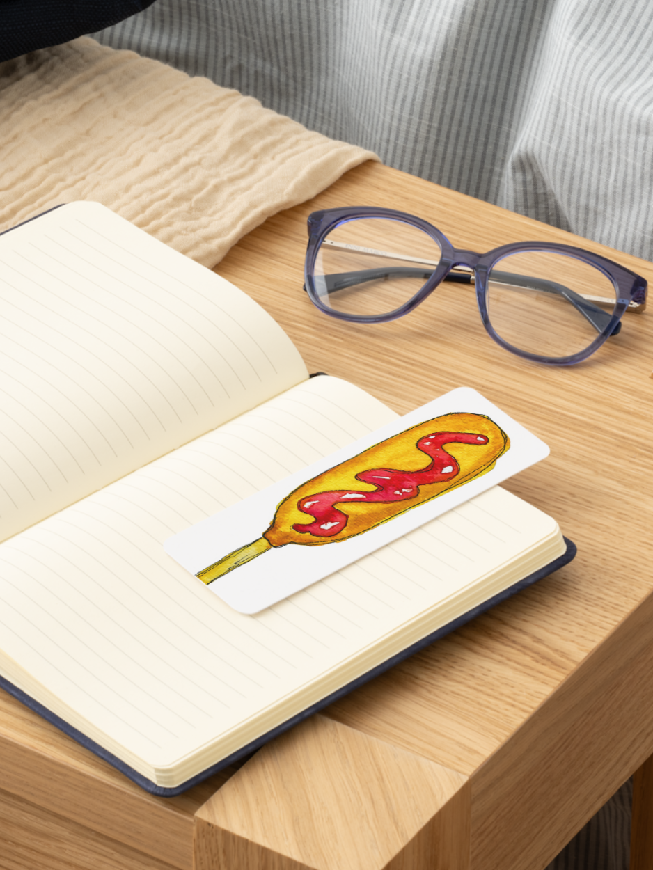 A rectangular white bookmark featuring a cartoon corn dog with ketchup, resting on an open lined notebook on a wooden table. Purple-framed eyeglasses and a beige blanket are also visible in the scene.