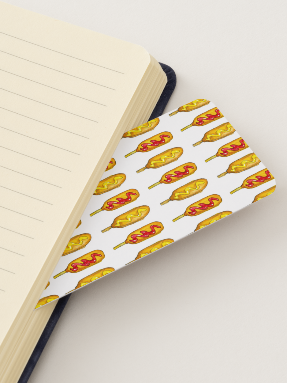 A close-up of a notebook with lined cream pages and a patterned bookmark or divider featuring illustrated corn dogs on a white background.