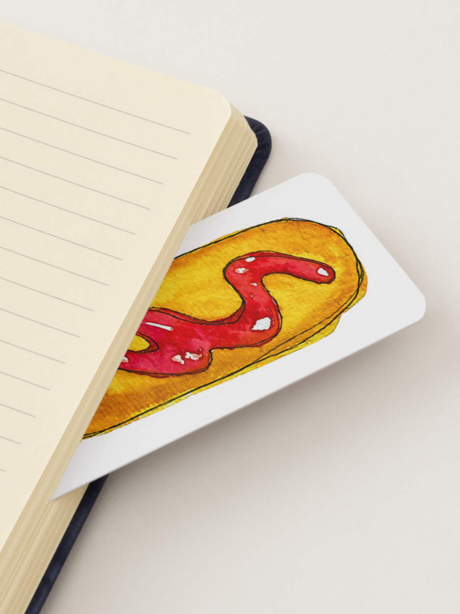 A white rectangular bookmark with rounded corners, featuring a watercolor illustration of a corn dog with red ketchup, resting on top of an open cream-colored lined notebook.