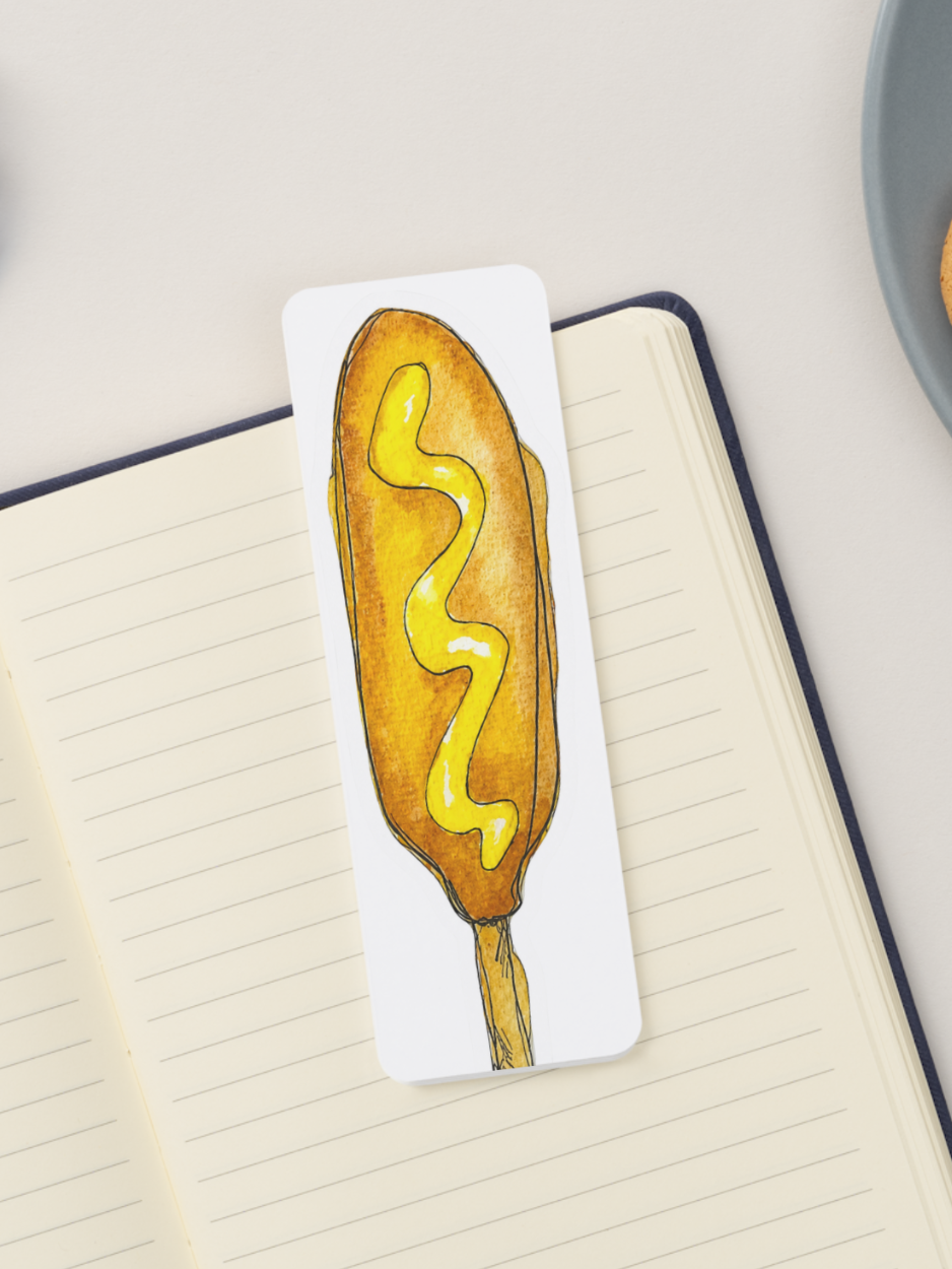 A white rectangular bookmark featuring a hand-drawn illustration of a corn dog with yellow mustard, placed on an open lined notebook.