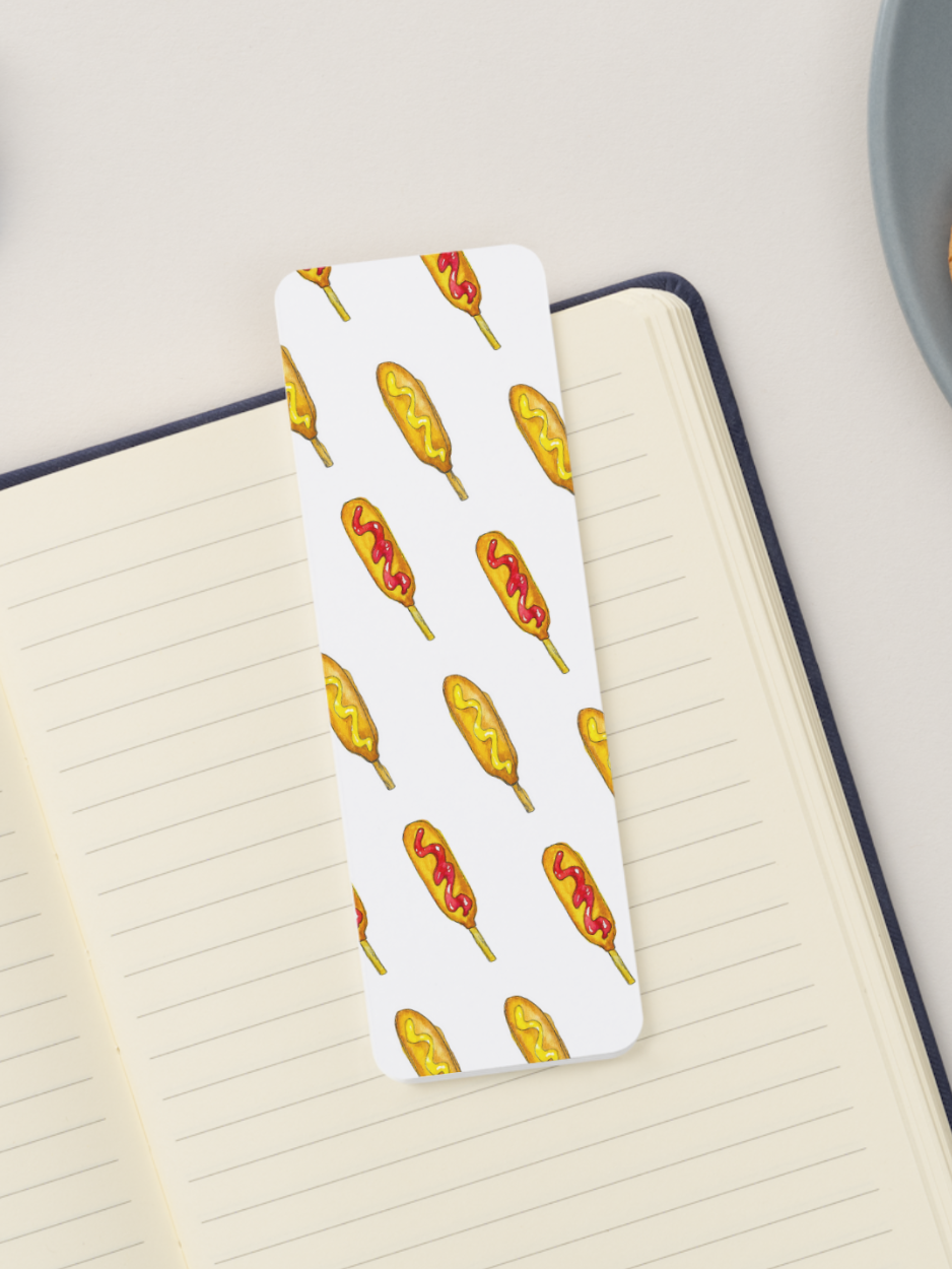 A white rectangular bookmark with rounded corners featuring a repeating pattern of illustrated corn dogs, placed on an open lined notebook.
