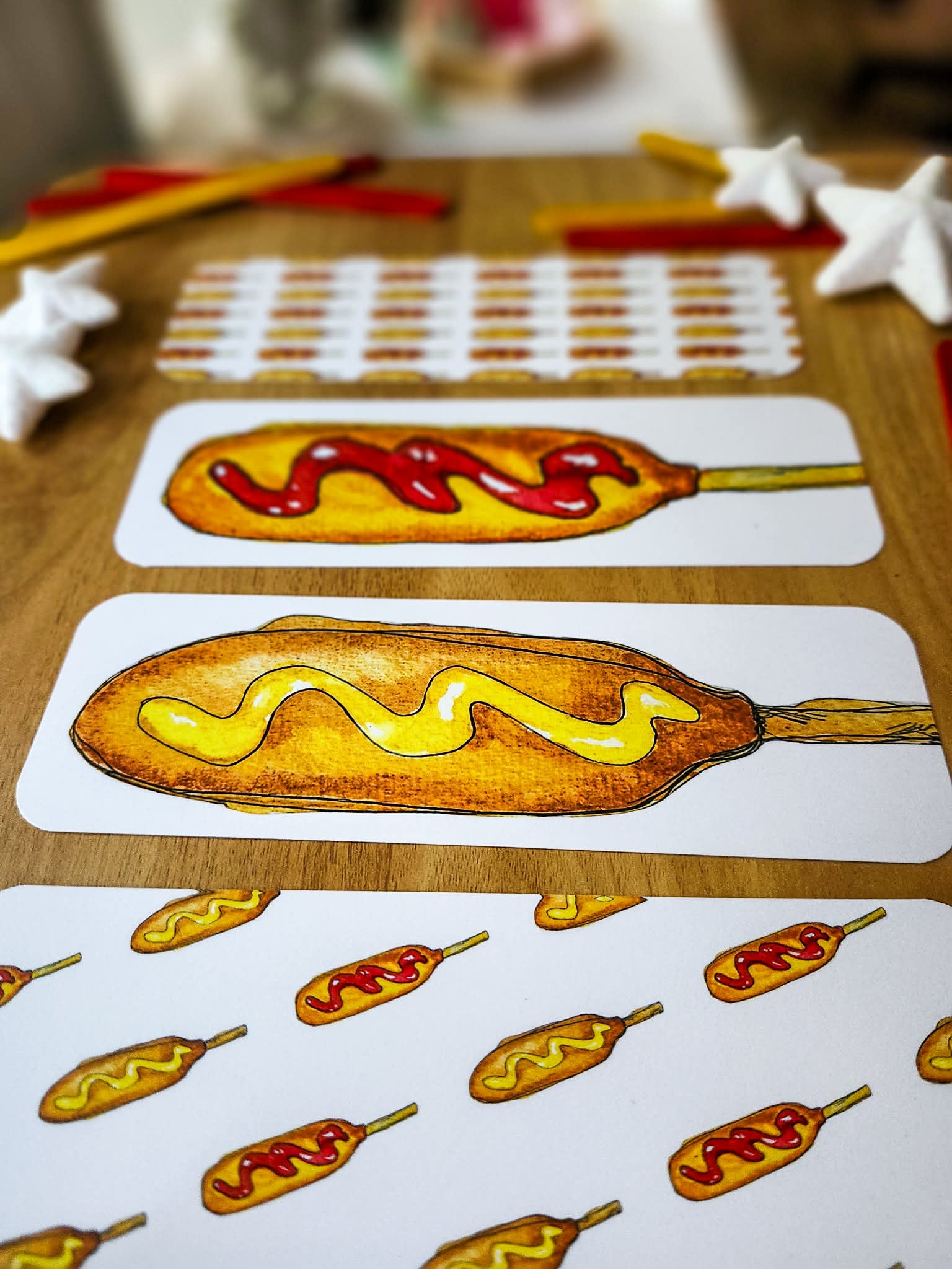 A close-up of hand painted watercolor bookmarks featuring illustrated corn dogs with mustard and ketchup, arranged on a wooden tabletop with small white star-shaped decorations and other craft-like items in the background.