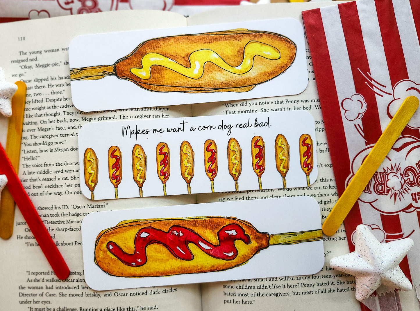 A set of illustrated bookmarks shaped like corn dogs with mustard and ketchup, displayed on top of an open book alongside red and white striped paper and star-shaped clips. Makes me want a corn dog real bad