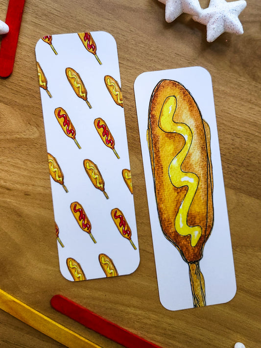 Two white rectangular bookmarks with corn dog illustrations, placed on a wooden surface. One card shows a repeating pattern of corn dogs with red mustard, and the other shows a single illustrated corn dog with yellow mustard.