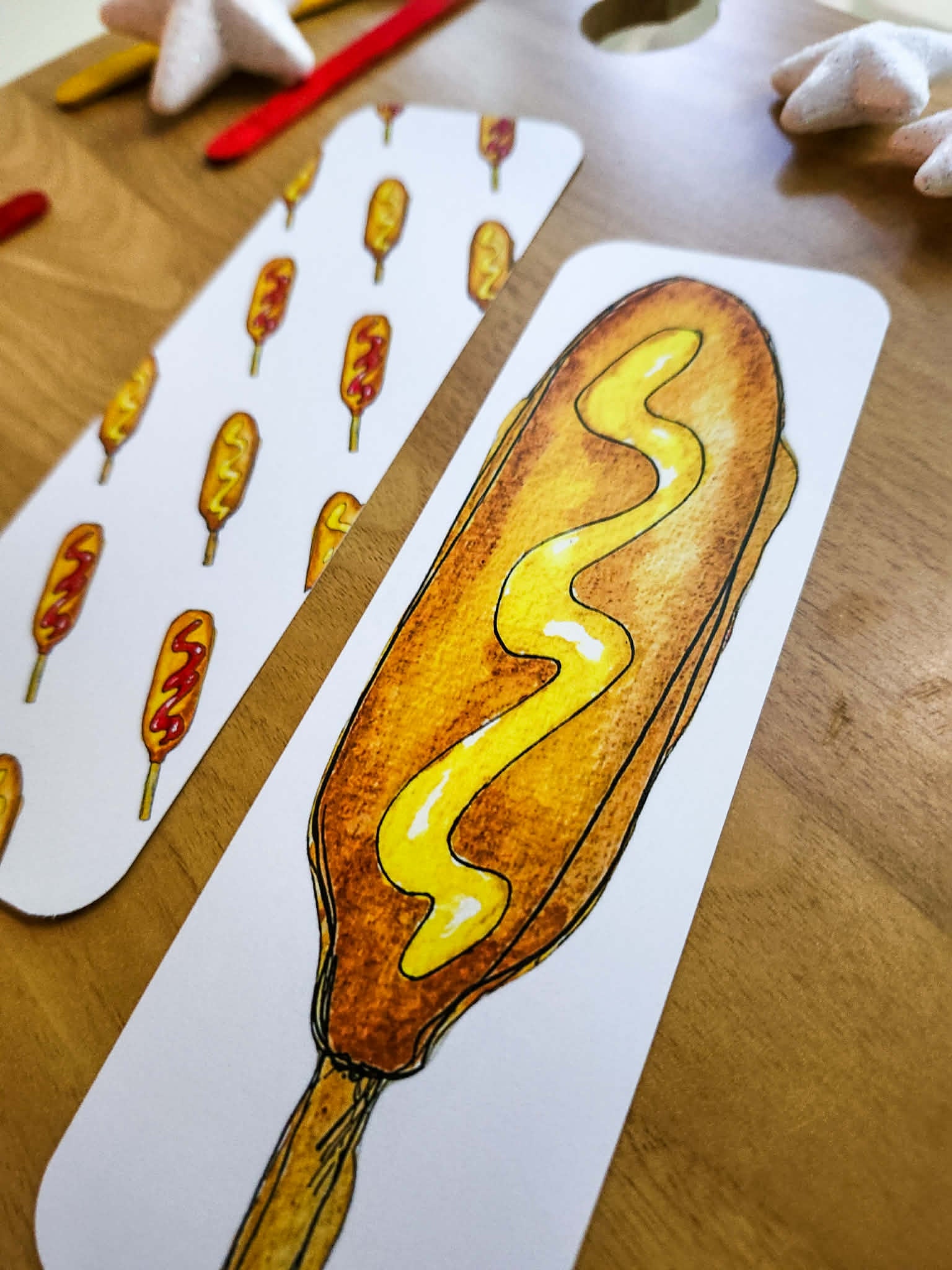 A close-up of printed bookmarks on a wooden table, showing a hand-drawn corn dog with mustard and a sheet with repeated corn dog illustrations.
