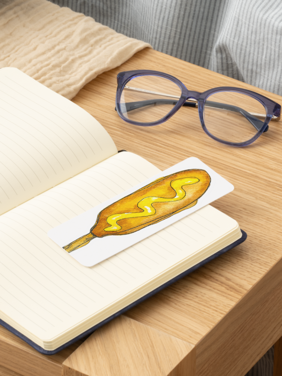 A rectangular white bookmark or card featuring a hand-drawn yellow mustard-covered corn dog, resting on an open lined notebook on a wooden table. Purple-framed eyeglasses and a beige knit blanket are visible in the background.