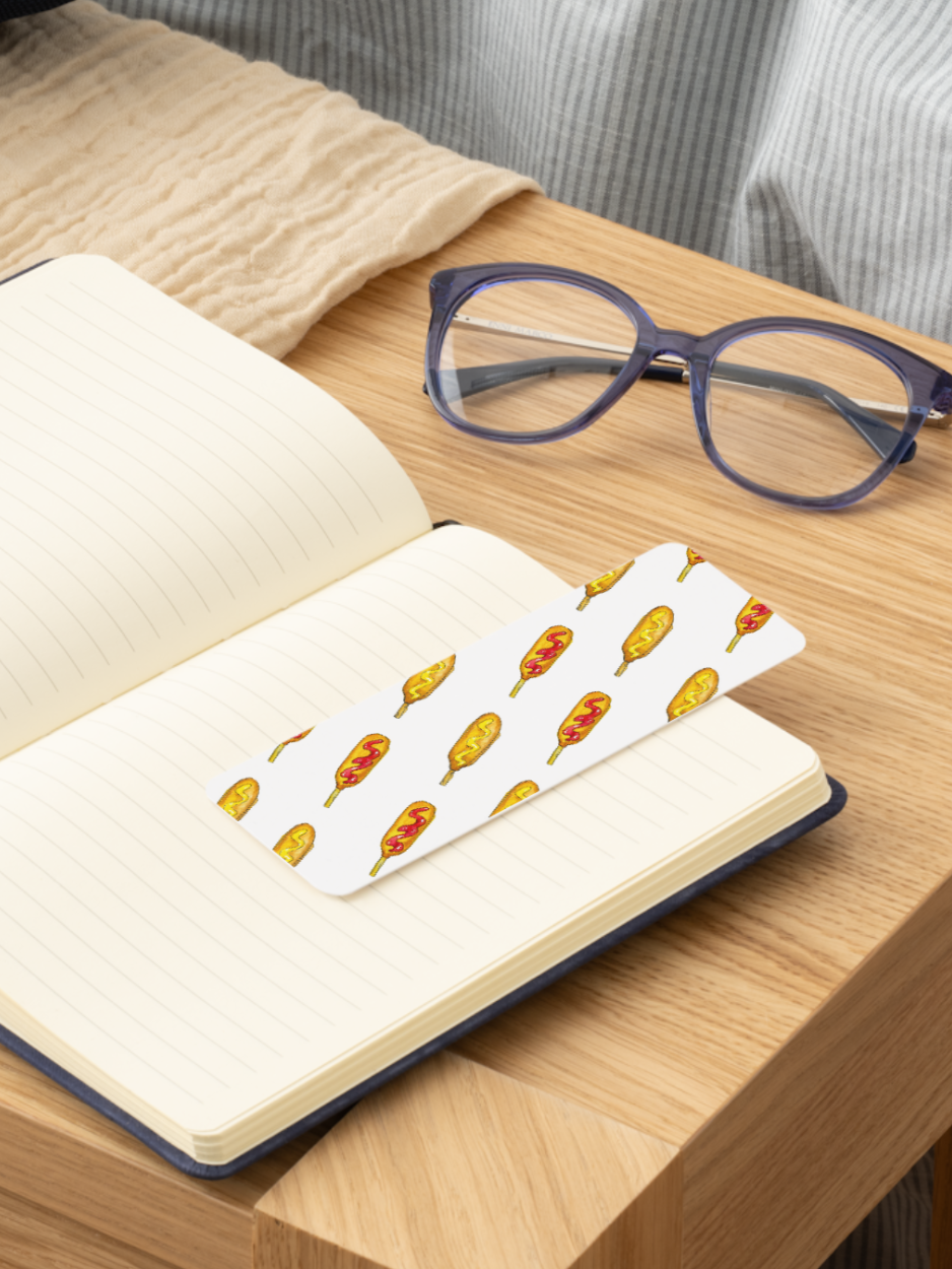 A white paper bookmark with a repeating pattern of corndogs, resting on the pages of an open notebook on a wooden table. A pair of dark purple eyeglasses is also on the table nearby.