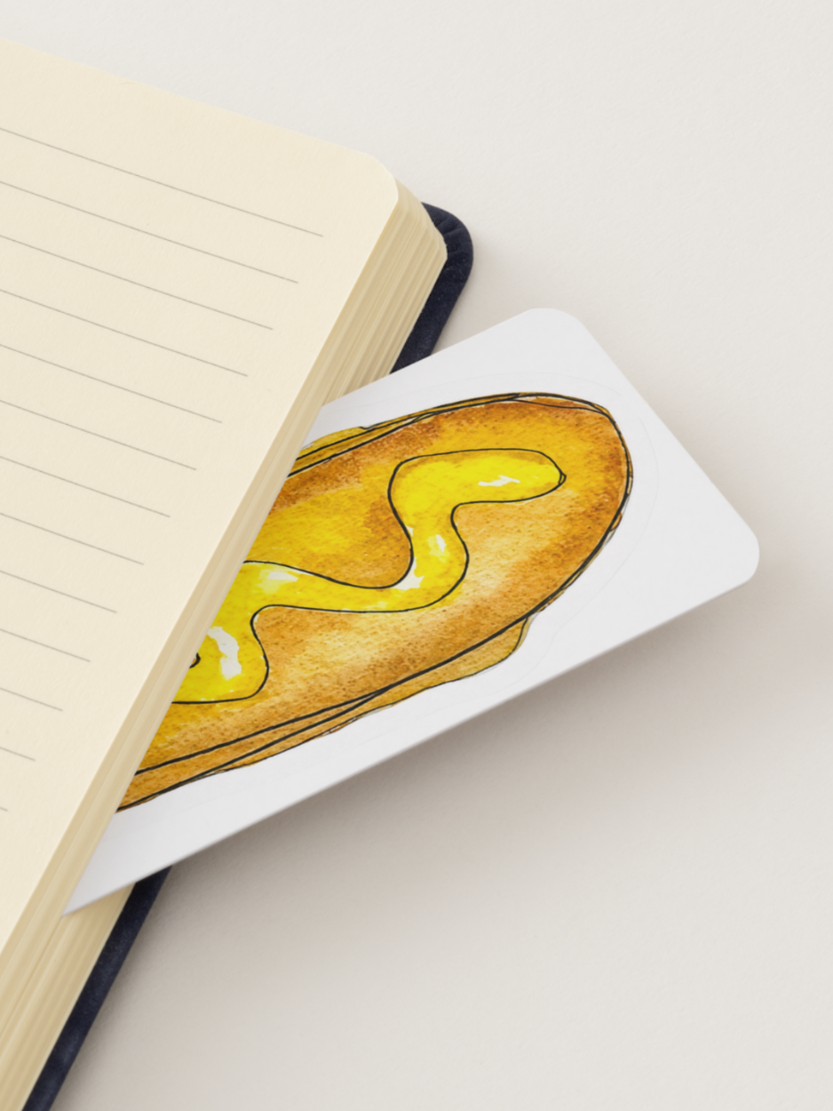 A white bookmark featuring a watercolor illustration of a toasted sandwich with yellow mustard, resting on a cream-colored lined notebook against a light beige background.