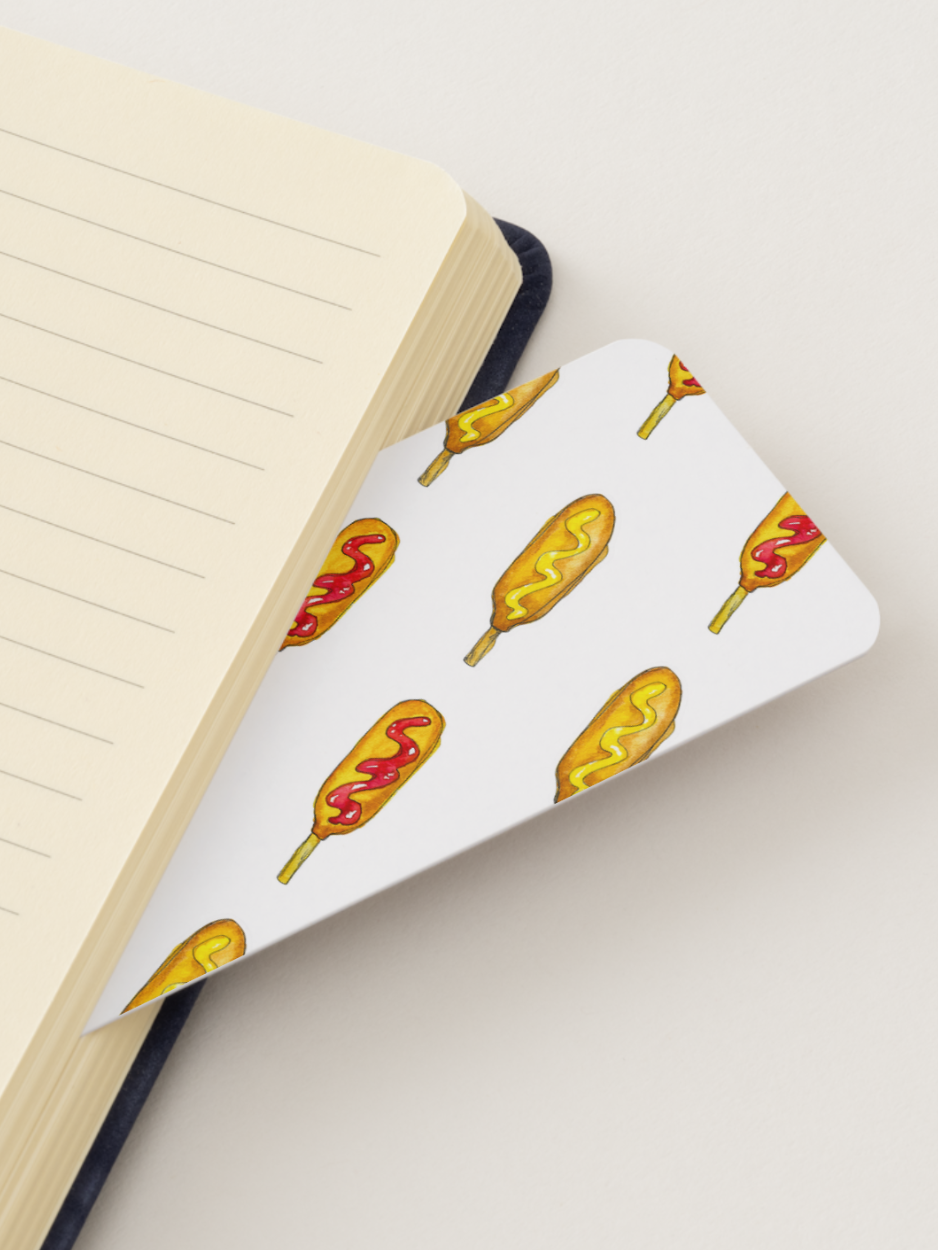 A white notebook bookmark with a repeating pattern of golden corn dogs with red ketchup, shown partially inserted under an open lined notebook on a light beige background.