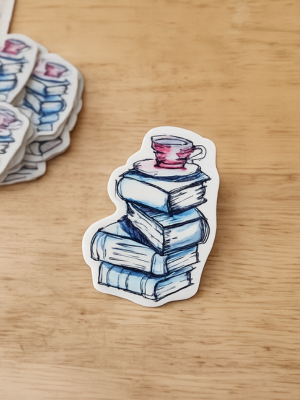 Sticker of a stack of books with a cup on top, placed on a wooden surface.