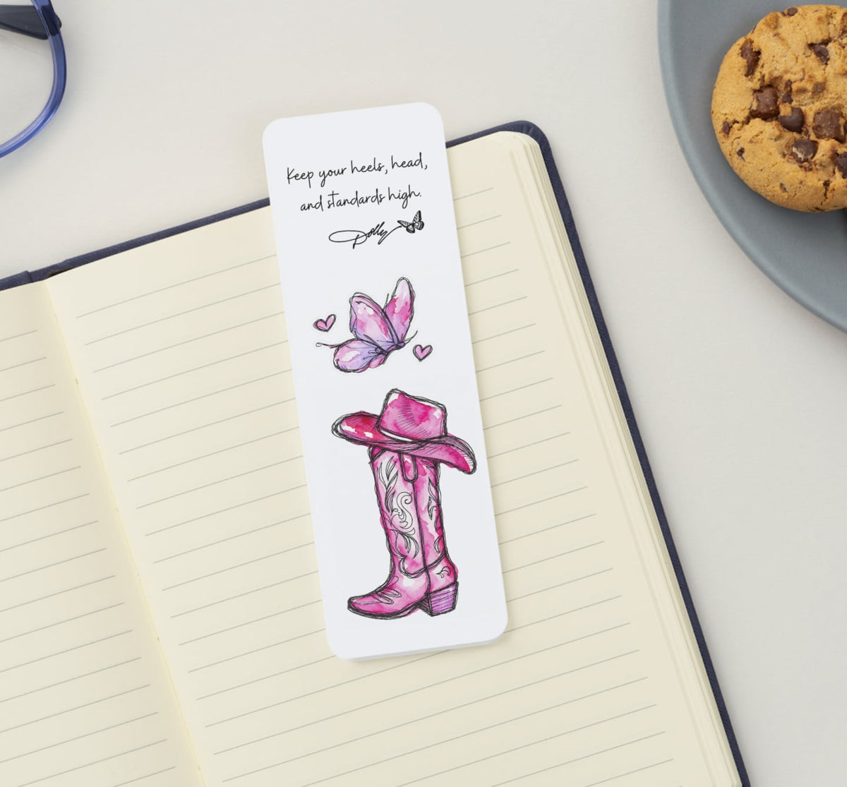Dolly Day 🦋🩷 | Limited Edition Watercolor Bookmark. A white rectangular bookmark with a pink cowboy boot and butterfly illustration, placed on an open lined notebook. The bookmark has printed text near the top and is photographed on a light surface with part of a cookie plate and eyeglasses visible in the background.