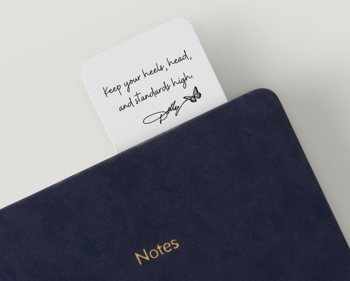 Dolly Day 🦋🩷 | Limited Edition Watercolor Bookmark. A navy blue textured notebook with gold 'Notes' lettering, shown open with a white card tucked inside. The card has black script text reading 'Keep your heels, head, and standards high.