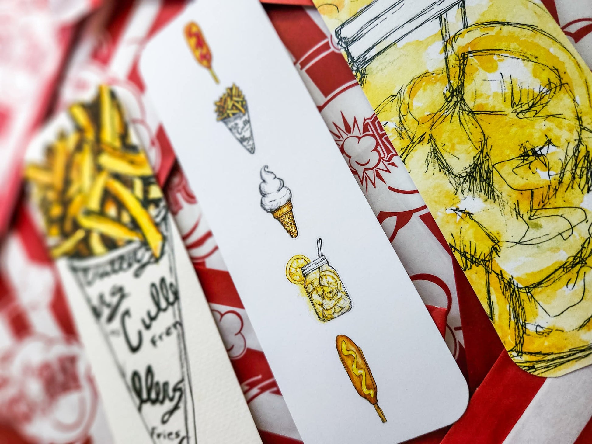 A close-up of several illustrated paper bookmarks laid on top of popcorn bags. The visible designs include Culvers french fries , an ice cream cone, a lemon shake up, and corndogs