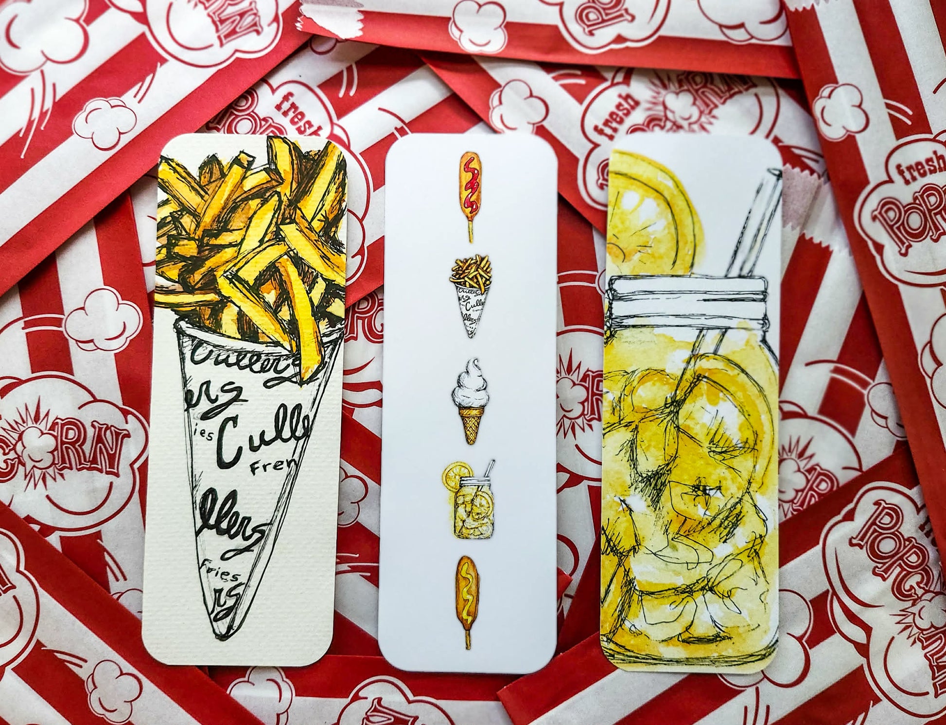 Three rectangular printed bookmarks laid on top of red-and-white popcorn bags. The left card shows illustrated french fries in a cone with handwritten text; the middle card shows small food illustrations in a vertical stack; the right card shows a mason jar filled with lemonade and a straw, with a lemon slice illustration above it.