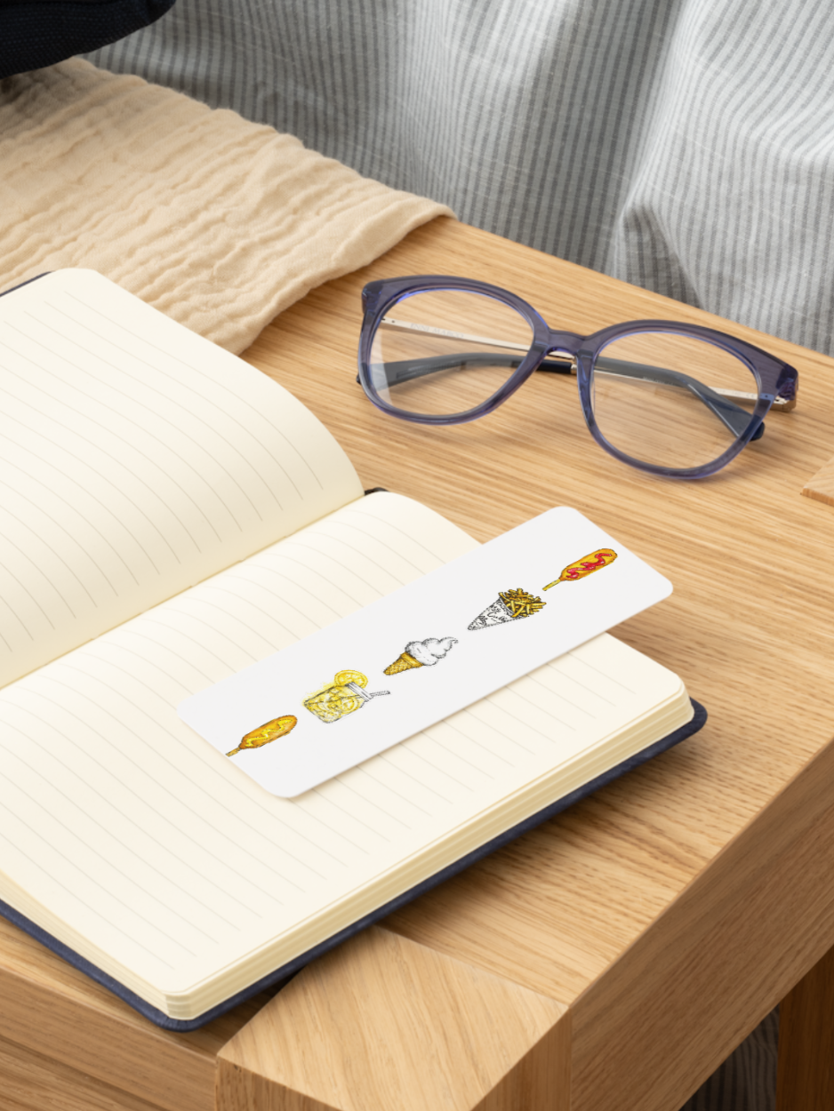 A white paper bookmark with cute illustrated characters lies across an open cream-colored lined notebook on a wooden table. A pair of purple-framed eyeglasses rests nearby, with a striped shirt and beige fabric in the background.