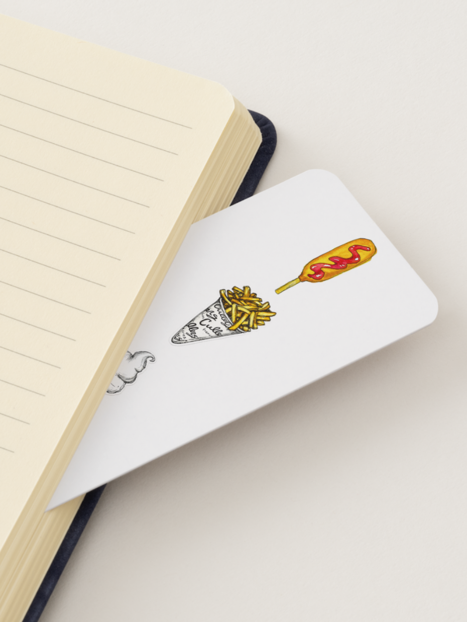 A white rectangular bookmark with a small illustration of french fries and a corn dog, partially tucked under the corner of an open cream-colored lined notebook on a light beige background.