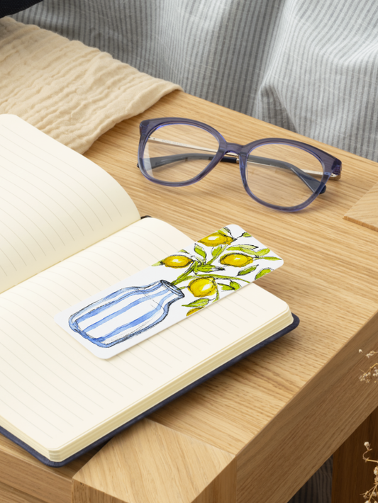 Open notebook with illustration bookmark of lemons and a bottle on a wooden surface with glasses in the background.