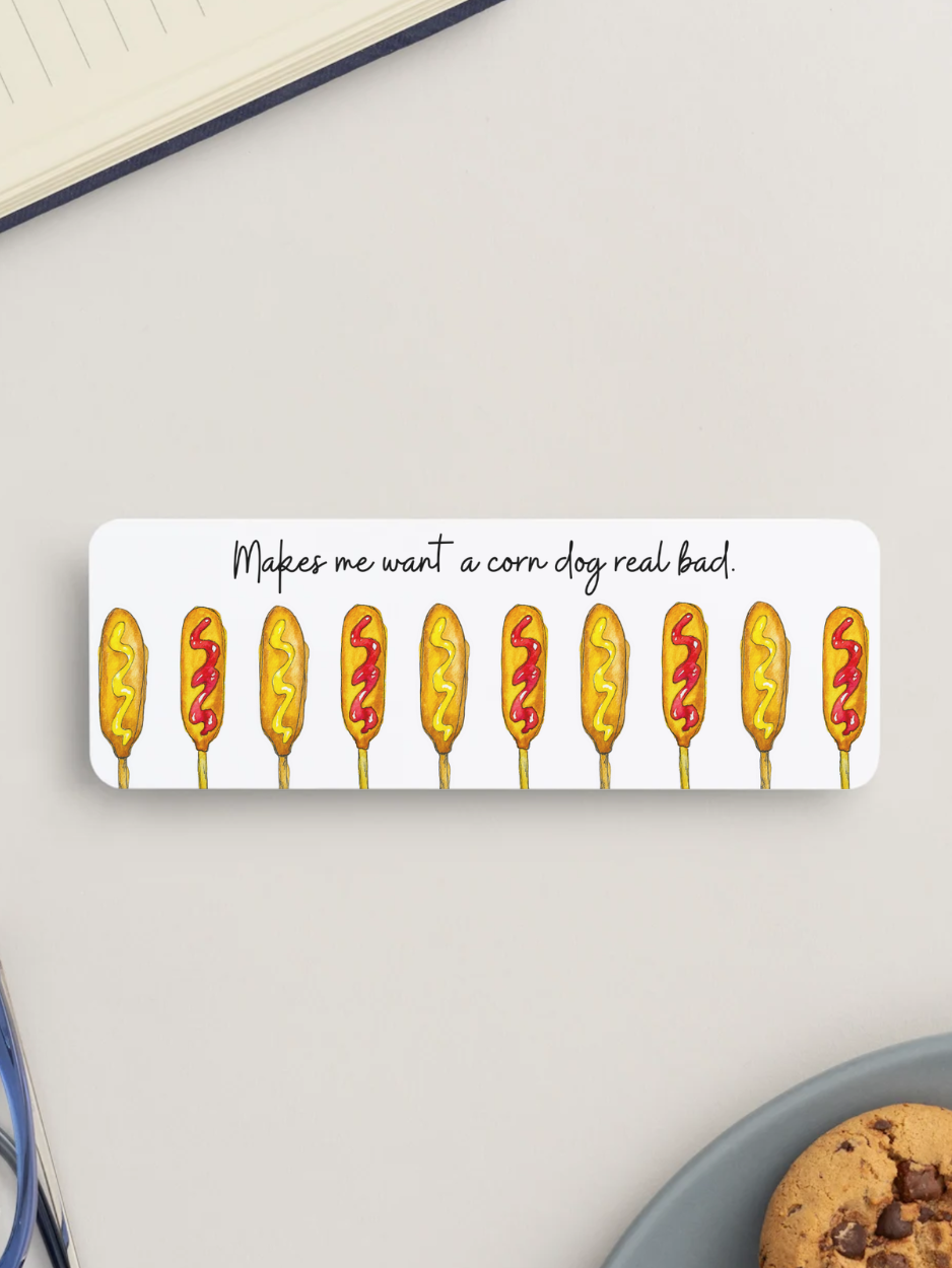 A white rectangular bookmark with rounded corners featuring a row of illustrated corn dogs and the text 'Makes me want a corn dog, real bad!' in black script, shown on a light gray surface with partial notebook and cookie visible around it.