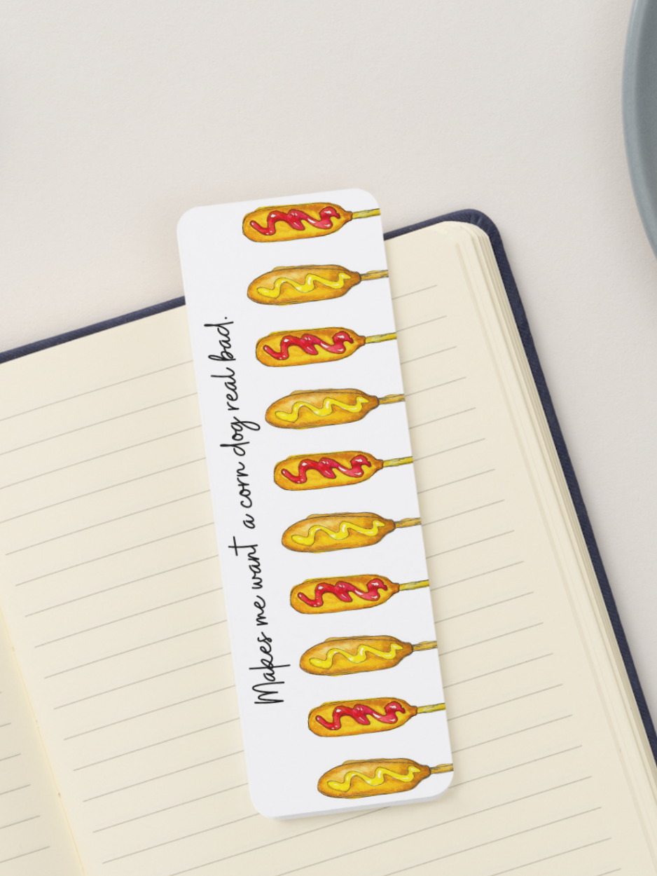 A white rectangular bookmark with a printed illustration of corn dogs and the phrase 'Makes me want a corn dog real bad.' placed on an open lined notebook.