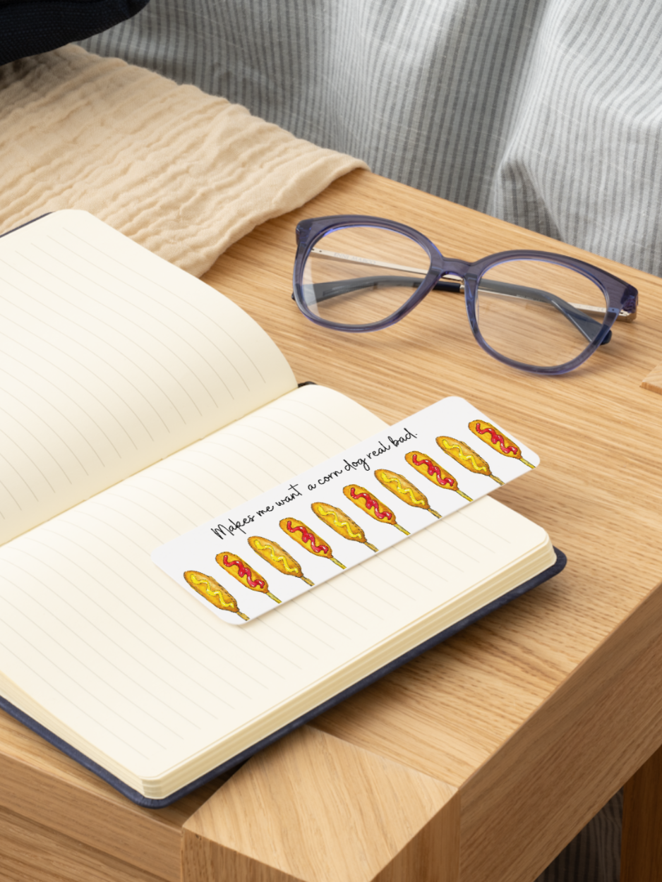 A white rectangular bookmark with printed corn dogs and the handwritten phrase 'Makes me want a corn dog real bad' rests on the pages of an open notebook on a wooden table. Purple-framed eyeglasses are also on the table nearby.
