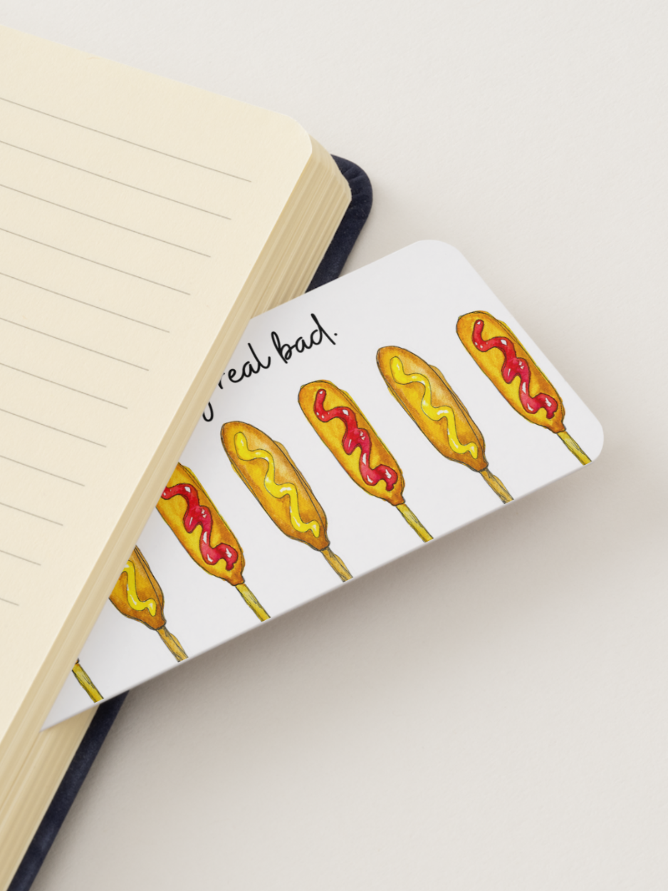 A white rectangular bookmark with illustrated corn dogs and the phrase 'Makes me want a corn dog real bad' placed over an open lined notebook on a light beige background.