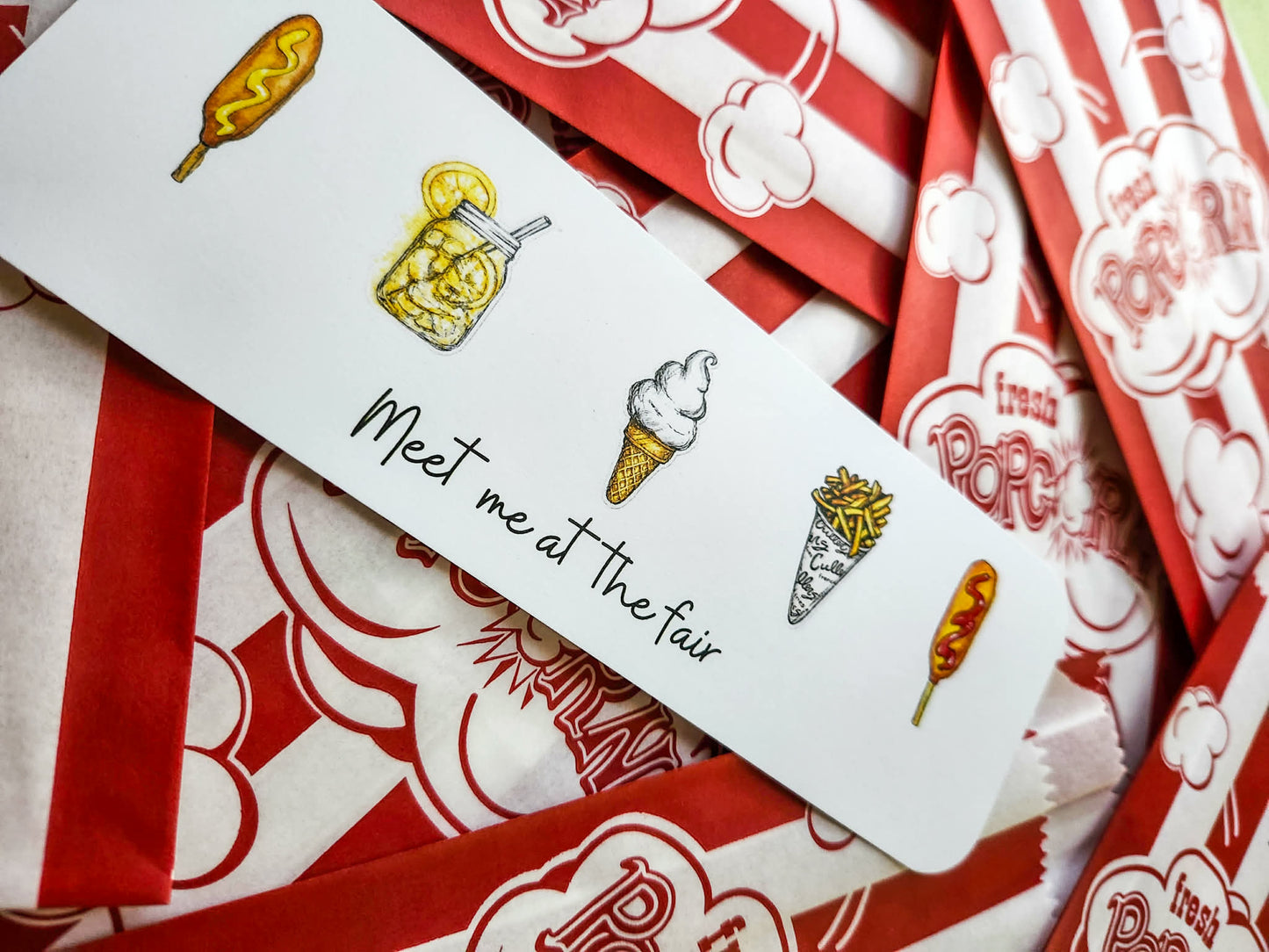 A white rectangular printed bookmark with hand-drawn food illustrations and the phrase 'Meet me at the fair' in black script, laid diagonally over red-and-white popcorn bags.