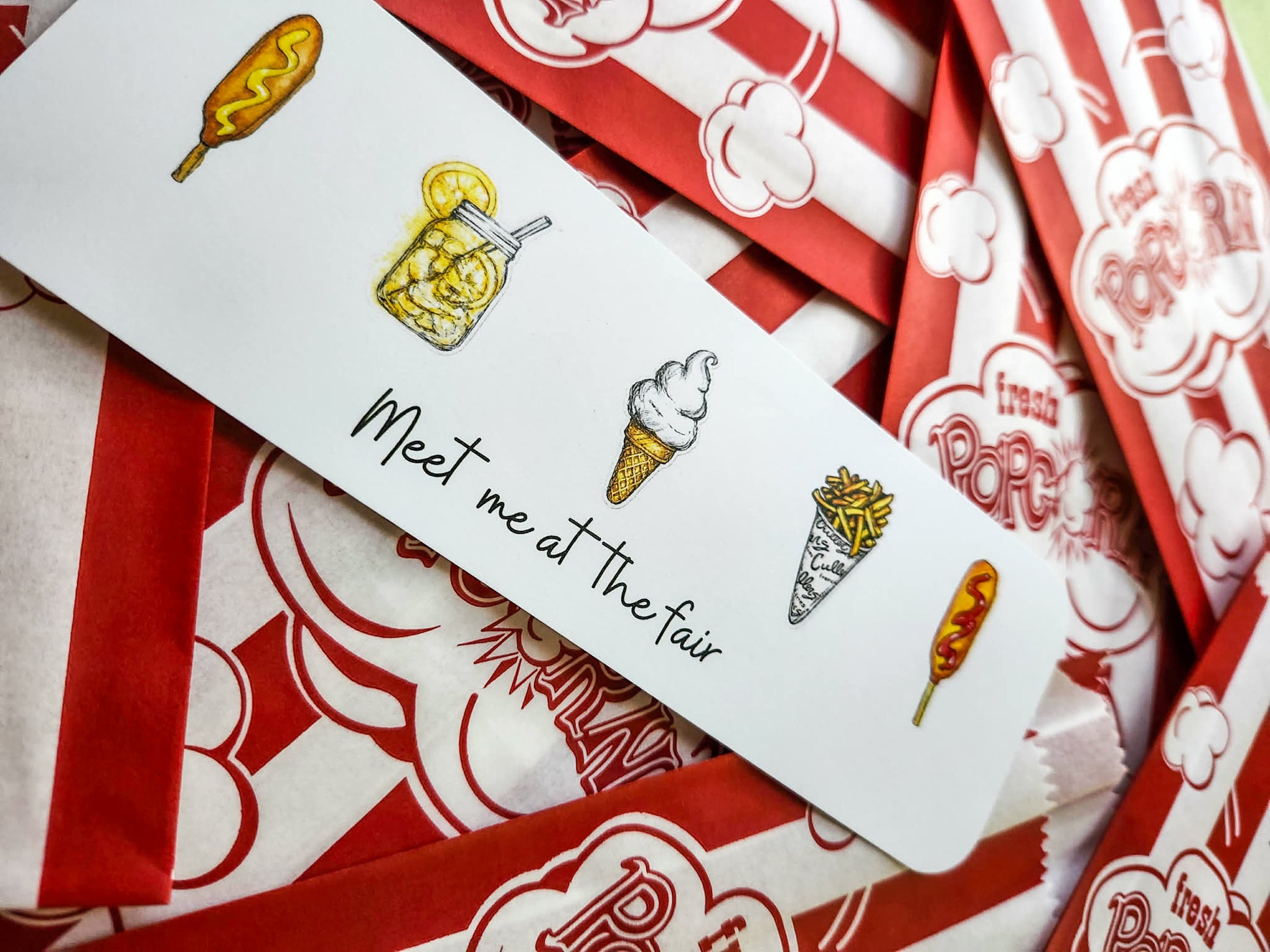A white rectangular printed bookmark with hand-drawn food illustrations and the phrase 'Meet me at the fair' in black script, laid diagonally over red-and-white popcorn bags.