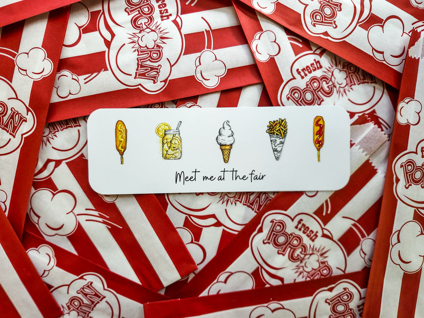 A white rectangular bookmark with illustrated snack icons and the text 'Meet me at the fair' lies on top of many red-and-white popcorn bags printed with 'Fresh Popcorn'.