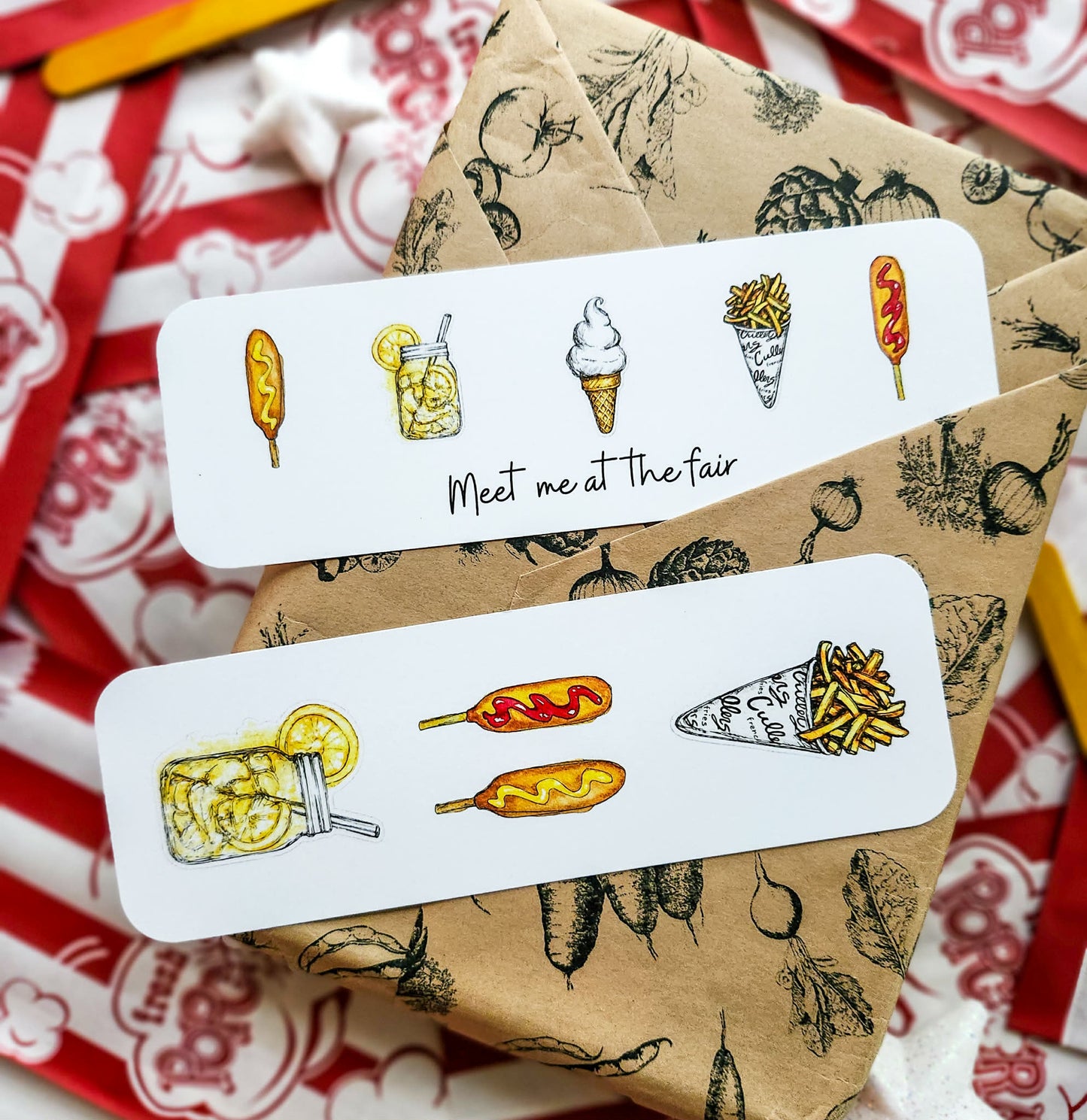 Two white rectangular bookmarks with hand-drawn food illustrations and the phrase 'Meet me at the fair' are arranged on top of brown patterned paper and red-and-white patterned fabric.