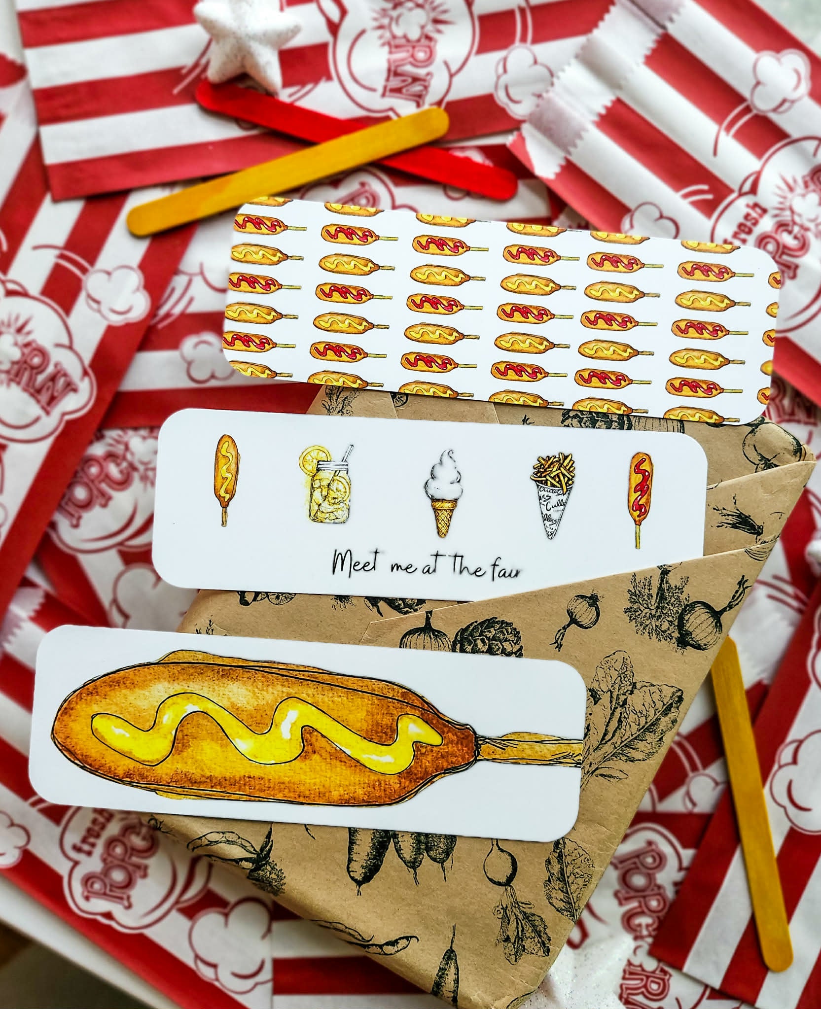 A set of food-themed bookmarks featuring illustrations of hot dogs, corn dogs, ice cream cones, and a drink, arranged on brown kraft paper and red-and-white patterned wrapping paper with popsicle sticks.