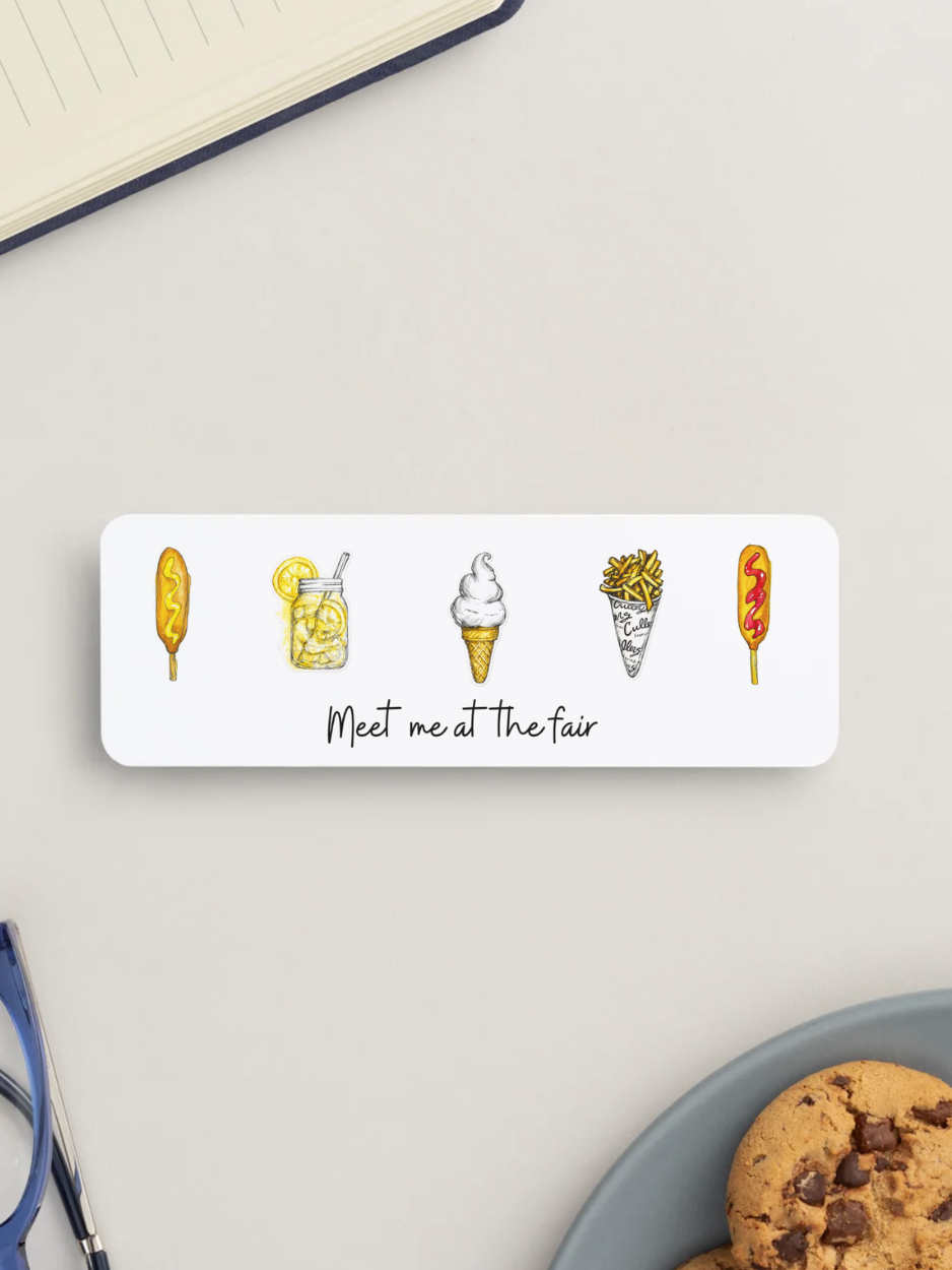 A white rectangular bookmark with rounded corners displayed on a light gray desk surface. The sticker features simple line-art illustrations of various treats and the handwritten text 'Meet me at the fair' in black script.