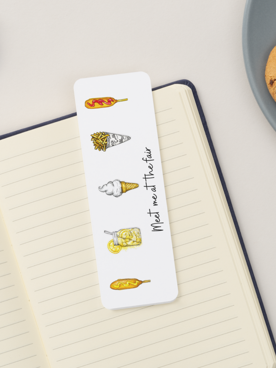 A white rectangular bookmark with illustrated food icons and the handwritten phrase 'Meet me at the fair' resting on an open lined notebook.
