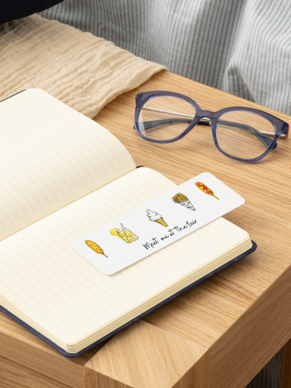 A white paper bookmark with colorful ice cream illustrations and the text 'Meet me at the bar' resting on an open lined notebook on a wooden table, with a pair of blue-purple eyeglasses nearby.