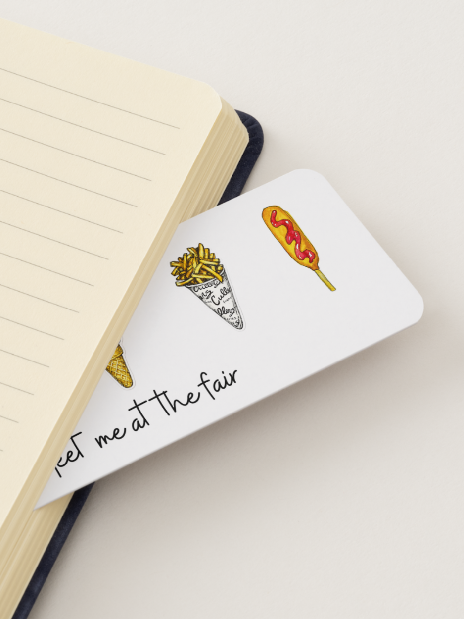 A white rectangular bookmark with illustrated food drawings and the partial text 'cket me at the fair' placed on top of an open cream-colored lined notebook.