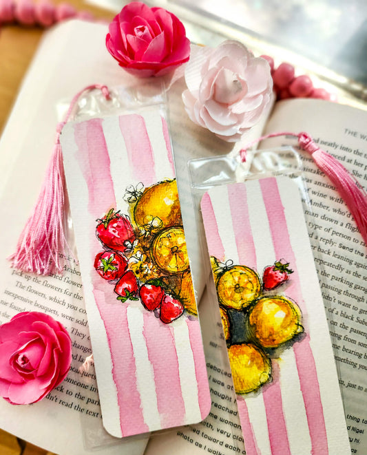 Two pink and white striped paper bookmarks with watercolor illustrations of lemons and strawberries, decorated with pink tassels, displayed on an open book with decorative pink roses.