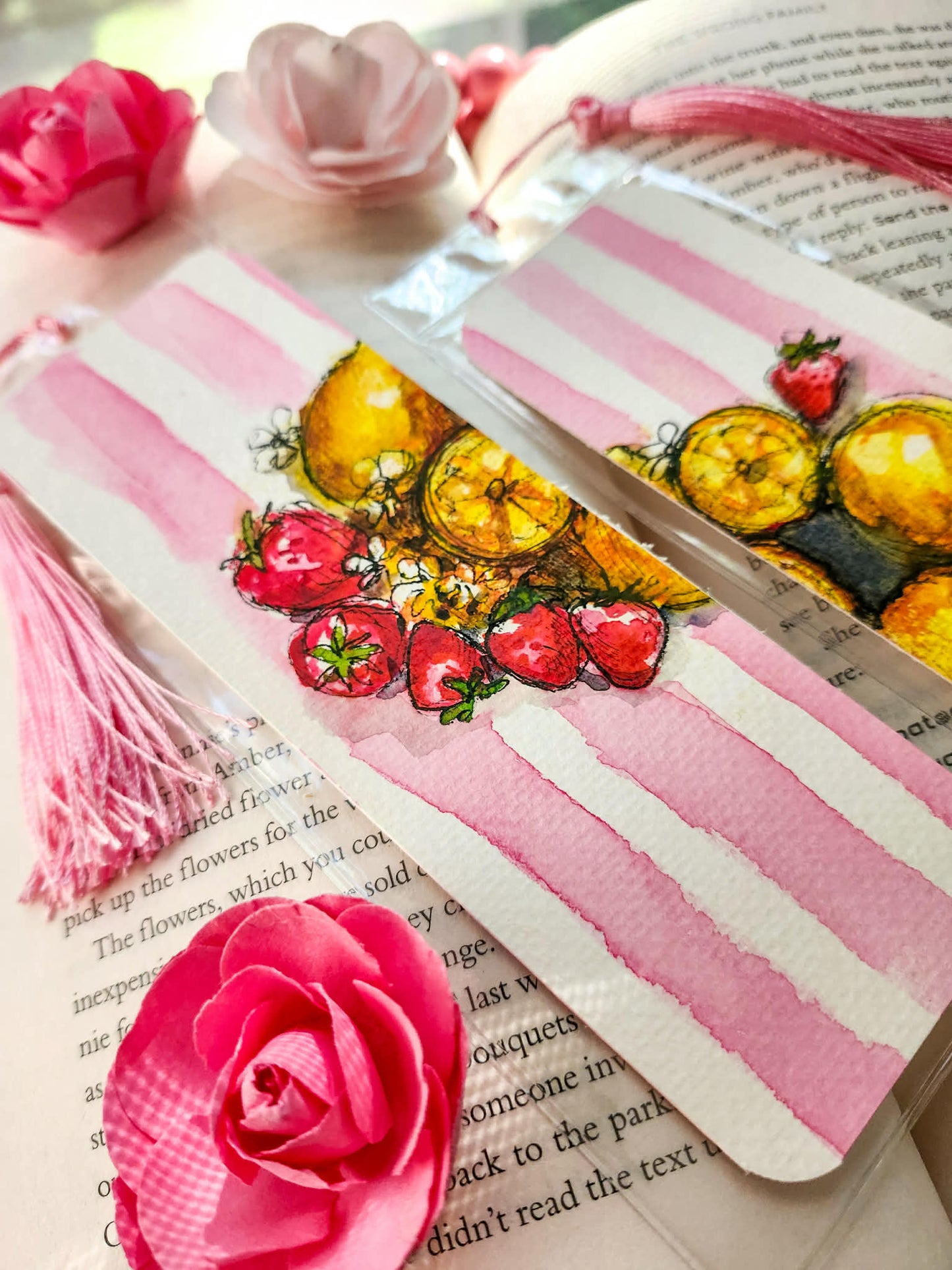 A close-up of two decorative bookmarks with pink watercolor stripes and painted fruit and strawberry motifs, placed on top of an open book with pink paper flowers and tassels around them.