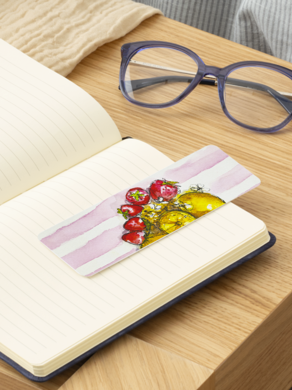 A rectangular bookmark with a watercolor illustration of red tomatoes and yellow olives on pink-and-white striped paper, placed on an open notebook on a wooden table. Purple eyeglasses and a cream knit fabric are also visible in the scene.