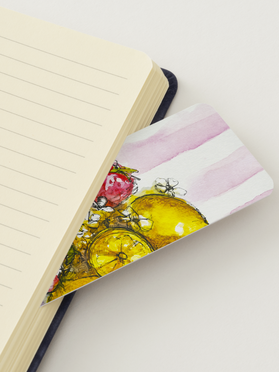A close-up of a lined notebook with an open page, resting on a slim notebook or card featuring a watercolor illustration of lemons, raspberries, and pink watercolor stripes.