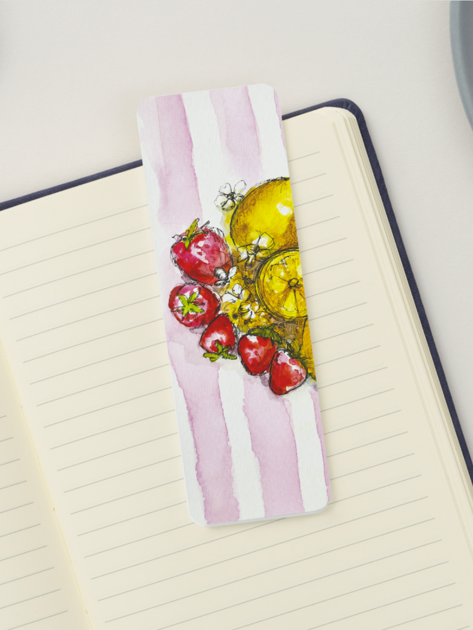 A rectangular watercolor bookmark with pink and white vertical stripes and an illustration of red strawberries and yellow citrus slices, placed on an open lined notebook.