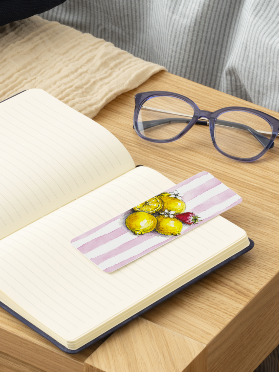 A close-up of an open notebook on a wooden table with a pink-and-white striped bookmark featuring yellow lemons and a strawberry. Purple eyeglasses rest nearby, with a beige blanket and gray striped fabric in the background.