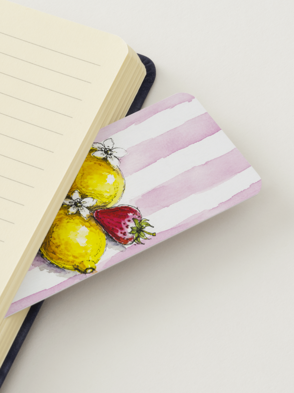A close-up of a notebook with cream-colored lined pages, partially covered by a decorative pink and white striped bookmark or notebook cover featuring watercolor illustrations of yellow lemons and a red strawberry.