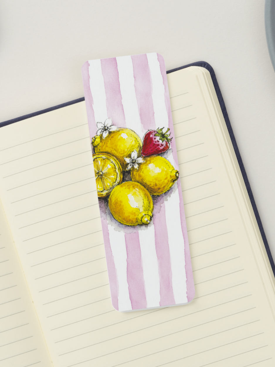 A rectangular paper bookmark with pink and white vertical stripes and an illustration of lemons and a strawberry, placed on an open lined notebook.