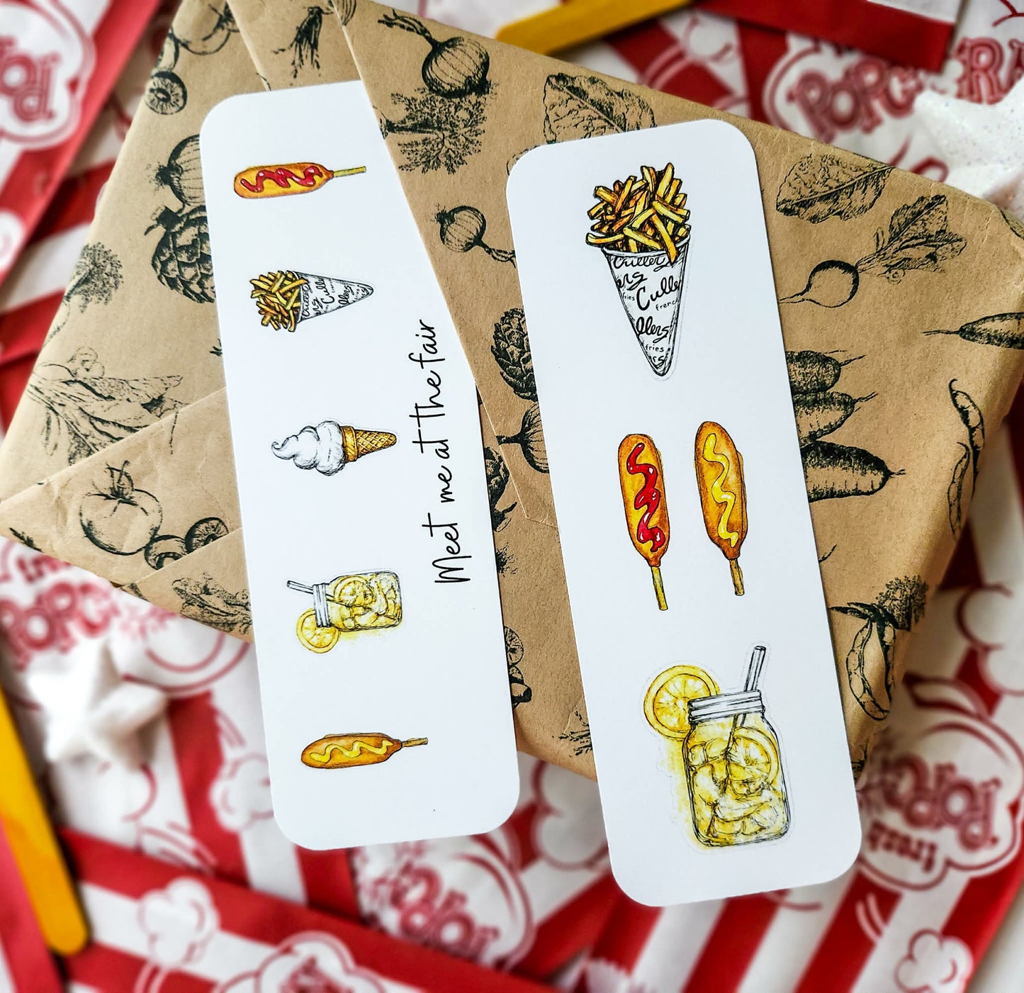 Two white rectangular bookmarks with illustrated food drawings and handwritten text, placed on top of brown patterned wrapping paper over red-and-white popcorn bags.