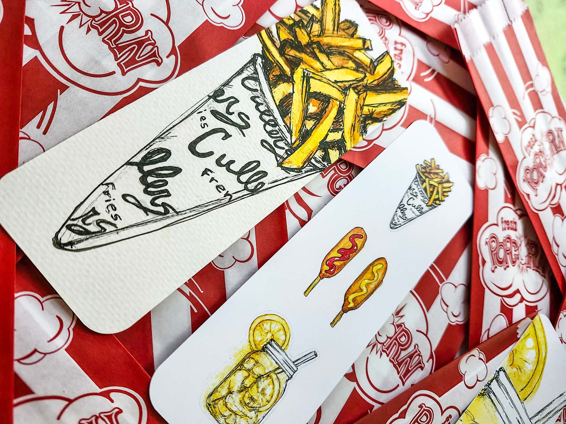 A close-up of illustrated bookmarks featuring french fries, corn dogs, a lemon shake up, and handwritten-style text, arranged on top of red-and-white popcorn bags