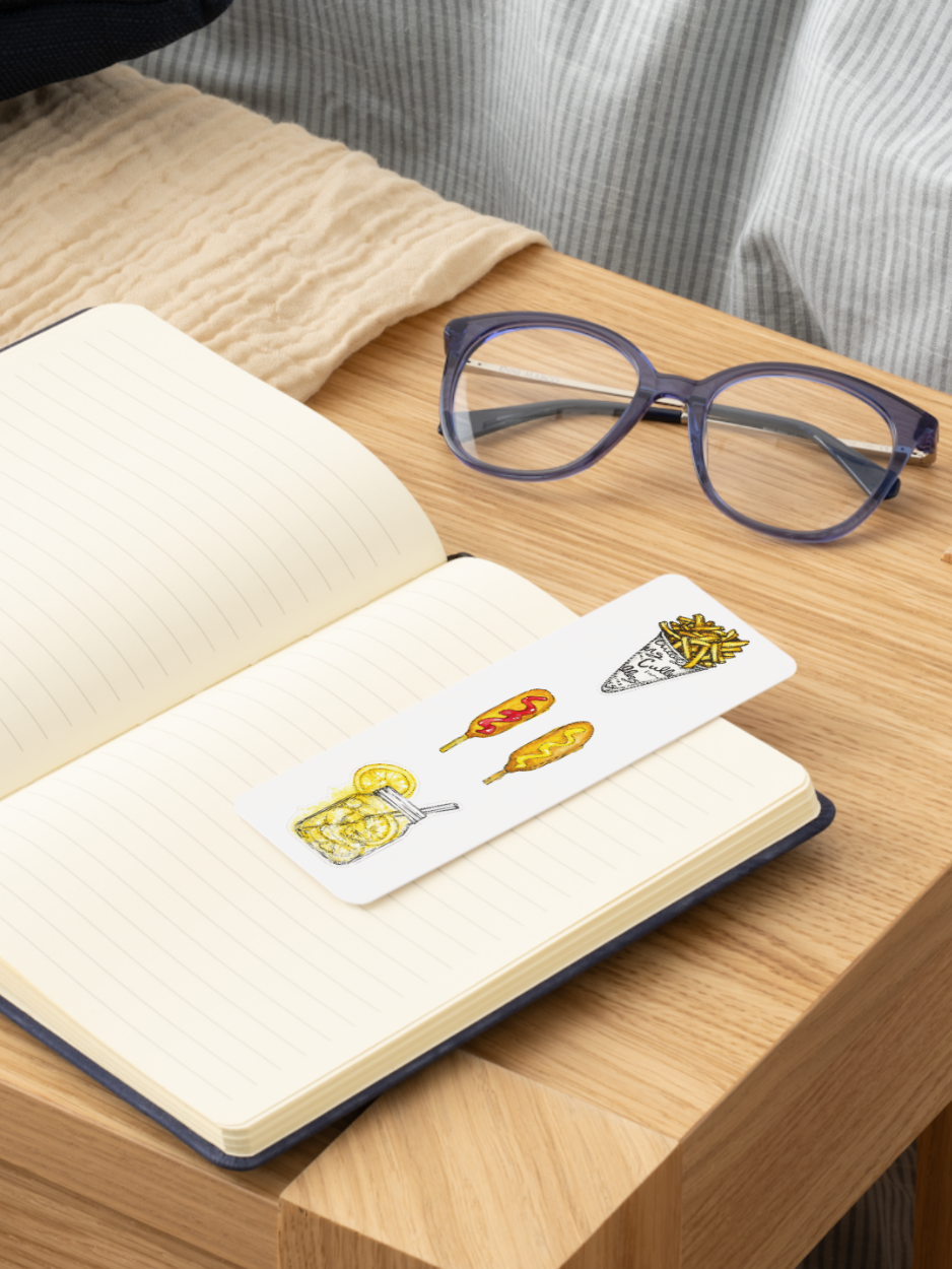 A white rectangular bookmark with colorful food illustrations rests on an open lined notebook on a wooden table, with a pair of eyeglasses nearby.